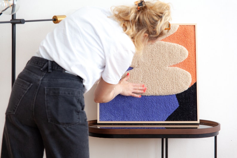 May include: A framed abstract wall art piece with a textured, multi-colored design. The artwork features a beige, orange, and purple color scheme with a black outline.