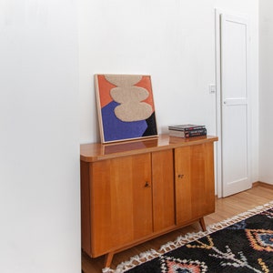 May include: A wooden cabinet with two doors, a framed abstract art print with beige, orange, blue, and black colors, and a stack of books on top. The cabinet is sitting on a wooden floor with a black and white rug with colorful accents.