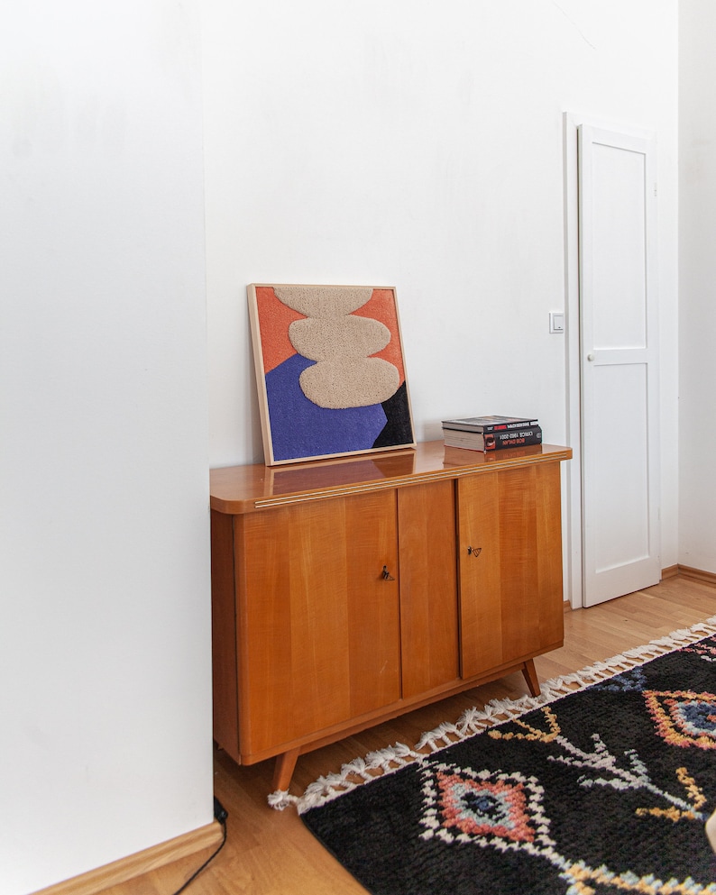 May include: A wooden cabinet with two doors, a framed abstract art print with beige, orange, blue, and black colors, and a stack of books on top. The cabinet is sitting on a wooden floor with a black and white rug with colorful accents.