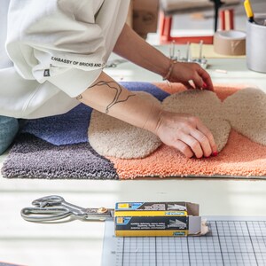 May include: A person is working on a colorful, abstract rug with a variety of textures. The rug is made of different shades of blue, beige, and orange. The person is using their hands to smooth out the rug. A pair of scissors and a box of staples are visible in the foreground.