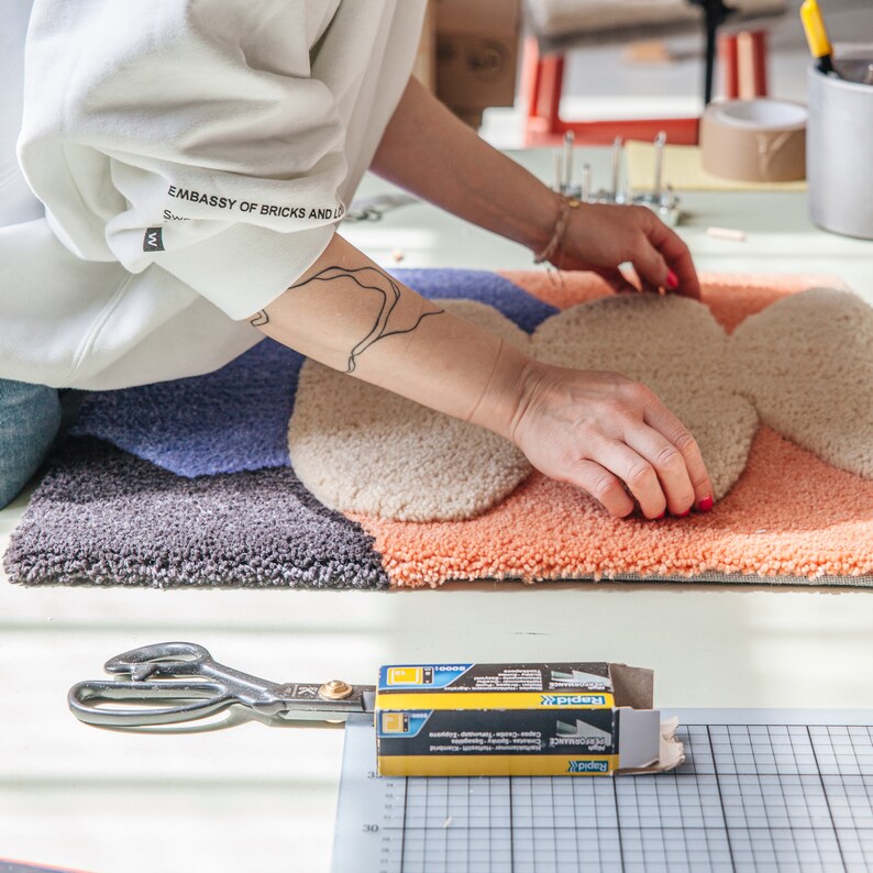 May include: A person is working on a colorful, abstract rug with a variety of textures. The rug is made of different shades of blue, beige, and orange. The person is using their hands to smooth out the rug. A pair of scissors and a box of staples are visible in the foreground.