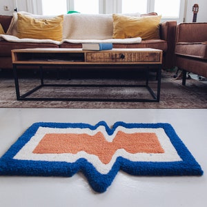Puede incluir: Una alfombra con estampado azul, blanco y naranja con un dise&ntilde;o ondulado. La alfombra est&aacute; sobre un suelo blanco delante de un sof&aacute; de cuero marr&oacute;n y una mesa de centro.