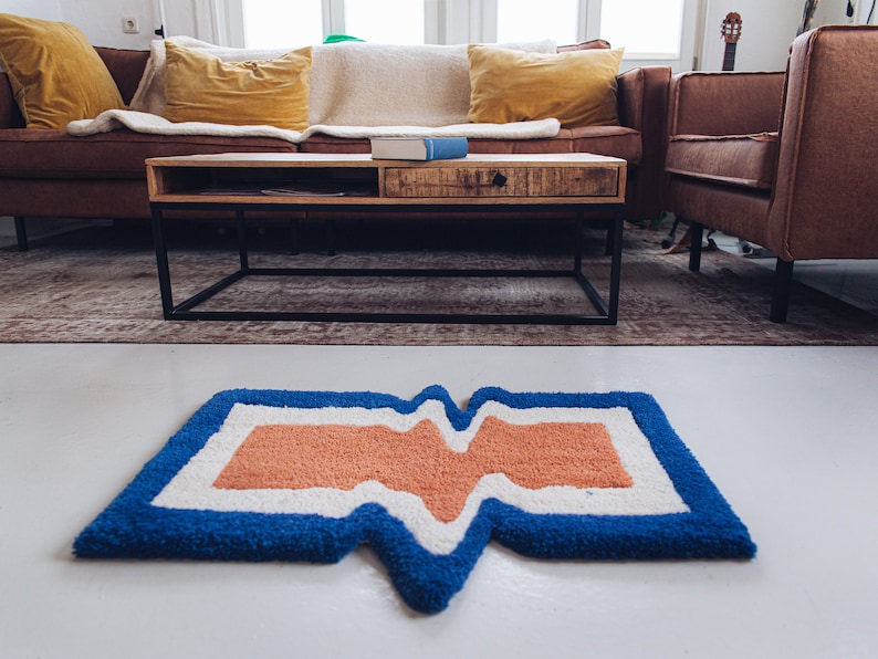Puede incluir: Una alfombra con estampado azul, blanco y naranja con un dise&ntilde;o ondulado. La alfombra est&aacute; sobre un suelo blanco delante de un sof&aacute; de cuero marr&oacute;n y una mesa de centro.
