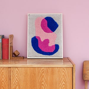 May include: A framed abstract wall art piece with a white frame and a gray background. The artwork features three overlapping shapes in shades of pink and blue.