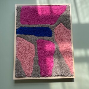 May include: A framed abstract wall art piece with a textured, fuzzy surface. The artwork features a variety of shapes in shades of pink, gray, and blue.