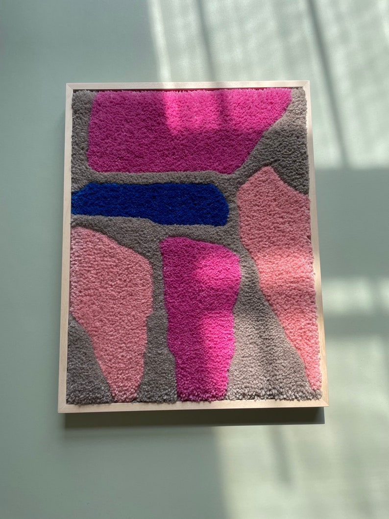 May include: A framed abstract wall art piece with a textured, fuzzy surface. The artwork features a variety of shapes in shades of pink, gray, and blue.