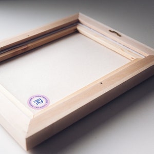 May include: A close-up of the back of a wooden canvas frame. The frame is light brown and has a white canvas stretched over it. A pink and blue sticker with the text "Colorfull Handmade Rosa Recuerdos" is on the bottom right corner of the frame.