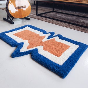 Puede incluir: Una alfombra abstracta azul, blanca y naranja con un dise&ntilde;o ondulado y org&aacute;nico. La alfombra est&aacute; hecha de un material suave y esponjoso.