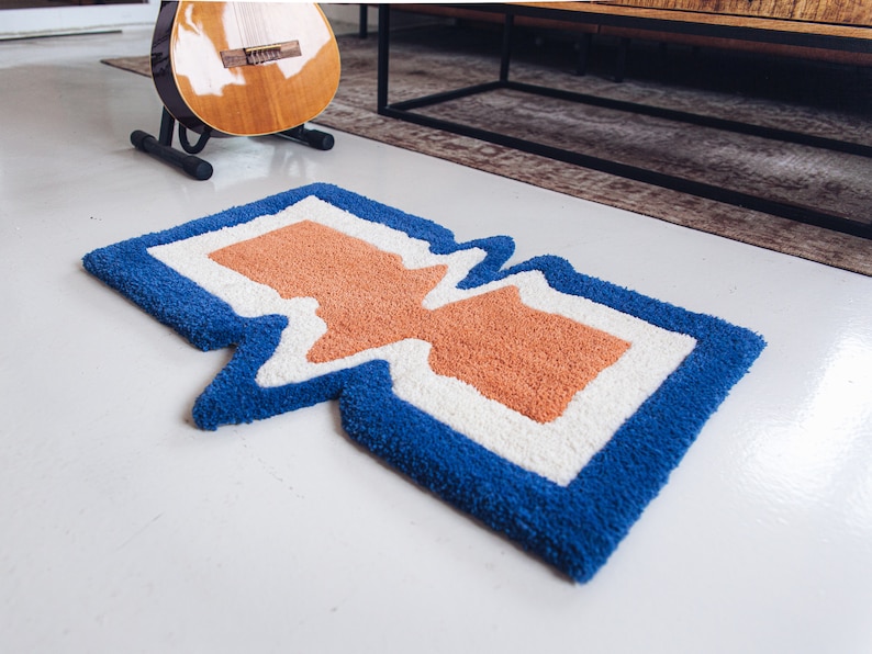Puede incluir: Una alfombra abstracta azul, blanca y naranja con un dise&ntilde;o ondulado y org&aacute;nico. La alfombra est&aacute; hecha de un material suave y esponjoso.