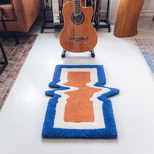 Puede incluir: Una alfombra azul, blanca y naranja con un dise&ntilde;o ondulado. La alfombra est&aacute; sobre un suelo blanco.