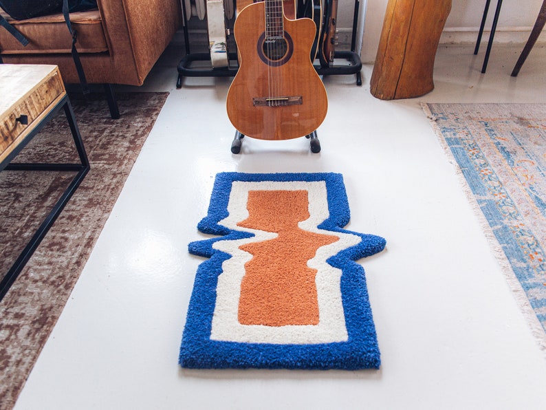 Puede incluir: Una alfombra azul, blanca y naranja con un dise&ntilde;o ondulado. La alfombra est&aacute; sobre un suelo blanco.