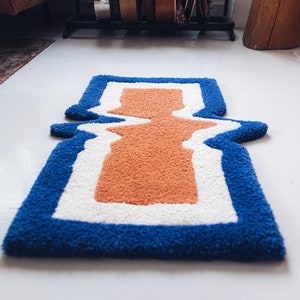 Puede incluir: Una alfombra azul, blanca y naranja con un dise&ntilde;o ondulado abstracto. La alfombra tiene una textura gruesa y esponjosa.