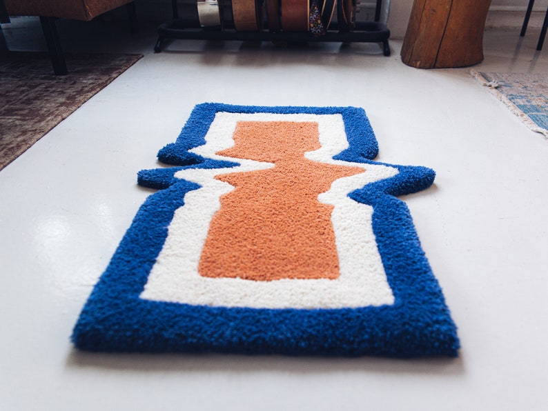 Puede incluir: Una alfombra azul, blanca y naranja con un dise&ntilde;o ondulado abstracto. La alfombra tiene una textura gruesa y esponjosa.