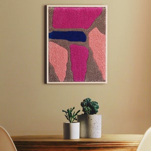 May include: A framed abstract wall art piece with pink, blue, and brown textured shapes. The art is hanging on a beige wall above a wooden table with two potted plants.