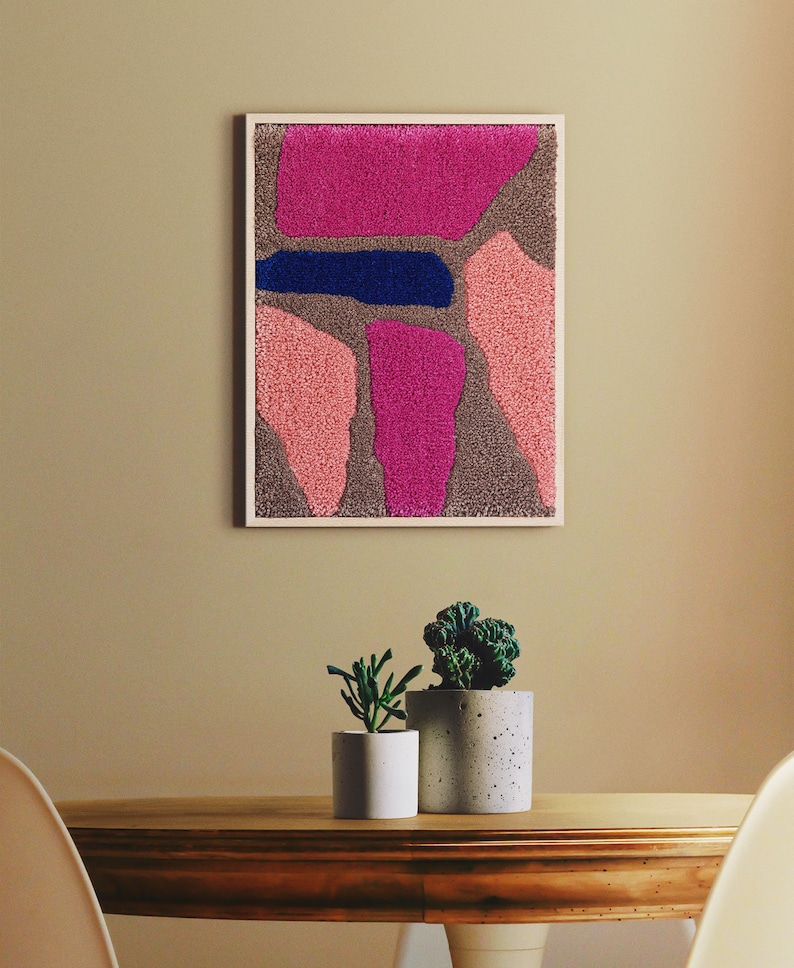 May include: A framed abstract wall art piece with pink, blue, and brown textured shapes. The art is hanging on a beige wall above a wooden table with two potted plants.