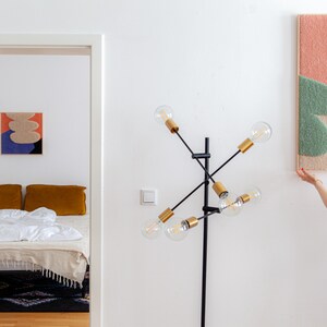 May include: A person hangs a colorful abstract wall tapestry with pink, green, and beige shapes on a white wall. The tapestry is made of yarn and has a textured surface.