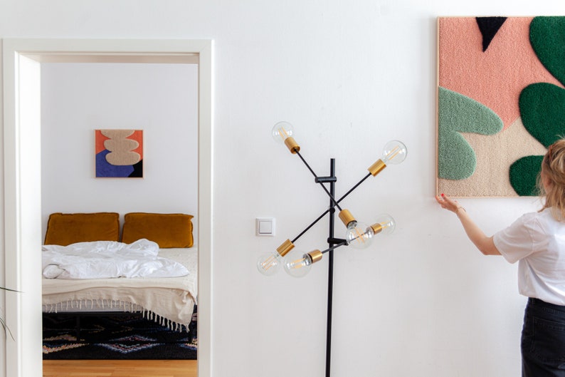 May include: A person hangs a colorful abstract wall tapestry with pink, green, and beige shapes on a white wall. The tapestry is made of yarn and has a textured surface.