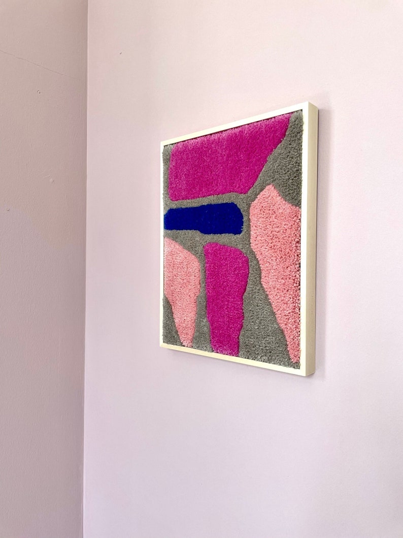 May include: A framed abstract wall art piece with a textured, fuzzy surface. The artwork features a variety of shapes in shades of pink, gray, and blue.
