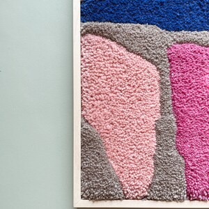 May include: A framed abstract wall art piece with a textured, fuzzy surface. The artwork features a variety of shapes and colors, including pink, blue, and gray.