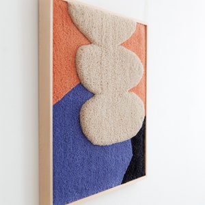 May include: A framed abstract wall art piece with a textured, fuzzy surface. The artwork features a layered design of three beige shapes on a background of orange, blue, and black.
