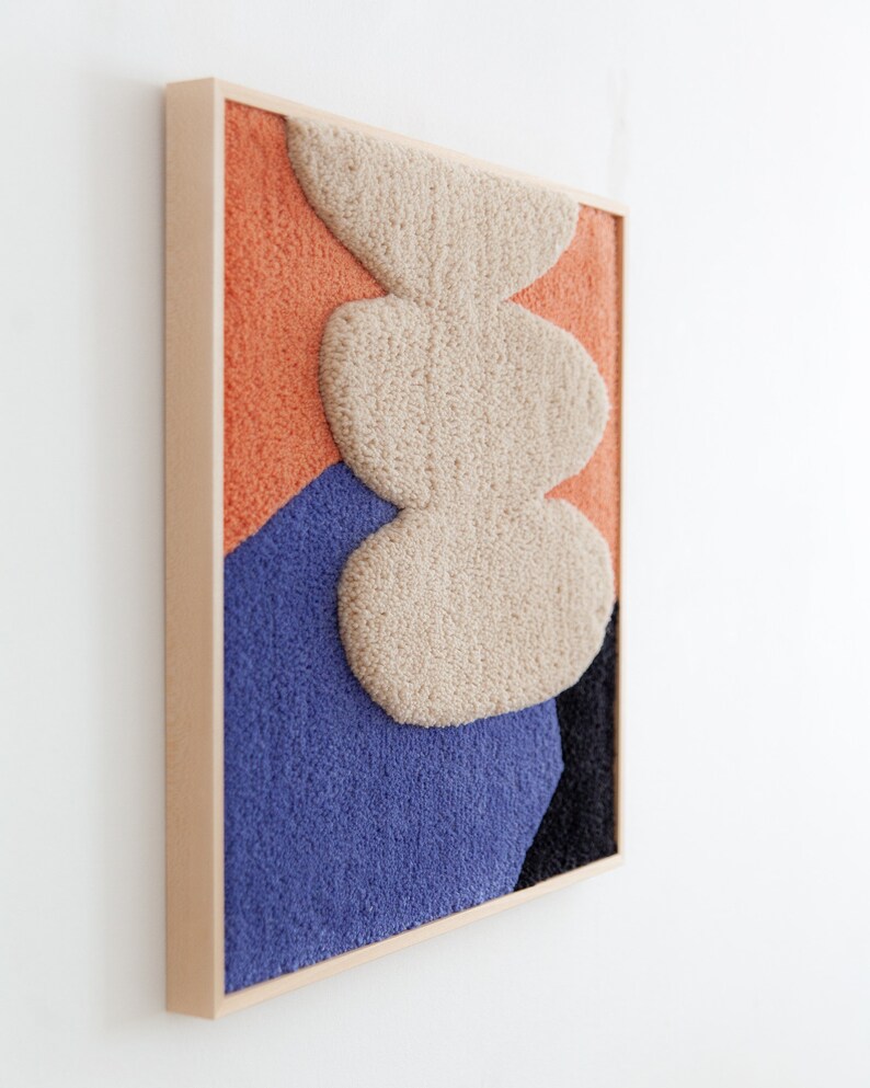 May include: A framed abstract wall art piece with a textured, fuzzy surface. The artwork features a layered design of three beige shapes on a background of orange, blue, and black.
