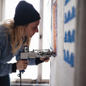 May include: A person using a tool to create a wall tapestry. The tapestry is made of blue and orange yarn and is being created on a white wall.