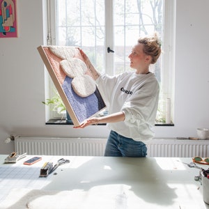 May include: A person holding up a framed abstract wall art piece. The art is made of colorful yarn and has a 3D effect. The frame is made of wood and has a natural finish.