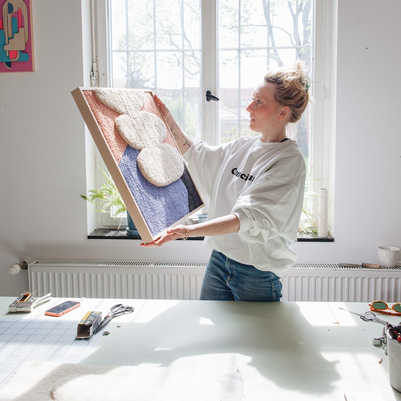 May include: A person holding up a framed abstract wall art piece. The art is made of colorful yarn and has a 3D effect. The frame is made of wood and has a natural finish.
