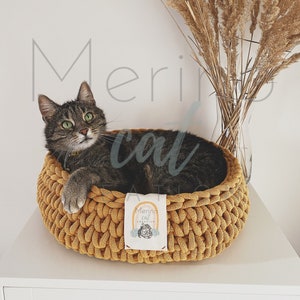 Puede incluir: Un gato atigrado descansando en una cama de gato de crochet de color mostaza. La cama tiene una etiqueta blanca con el texto "Merino Cat Creation" y un gr&aacute;fico de arco&iacute;ris.