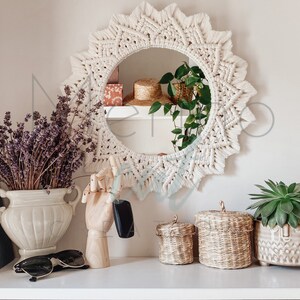 May include: A white macrame mirror with a sunburst design hangs on a white wall.  The mirror is surrounded by dried lavender, a wooden hand holding a car key, and three woven baskets.  Two of the baskets are on a shelf and one is on the floor.