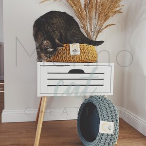 Puede incluir: Un gato sentado en una cama de gato de crochet amarillo en una mesita de noche blanca con una cama de gato de crochet azul en el suelo. La mesita de noche tiene patas de madera y un caj&oacute;n blanco con tres l&iacute;neas horizontales.