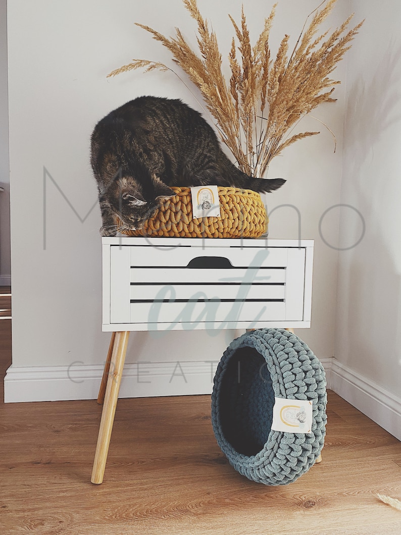 Puede incluir: Un gato sentado en una cama de gato de crochet amarillo en una mesita de noche blanca con una cama de gato de crochet azul en el suelo. La mesita de noche tiene patas de madera y un caj&oacute;n blanco con tres l&iacute;neas horizontales.