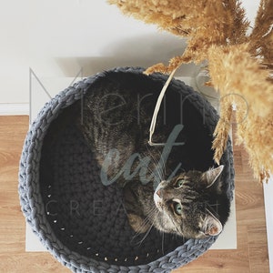 Puede incluir: Una cama para gatos de crochet gris con un gato atigrado descansando dentro. La cama para gatos es redonda y tiene un borde grueso de cuerda.