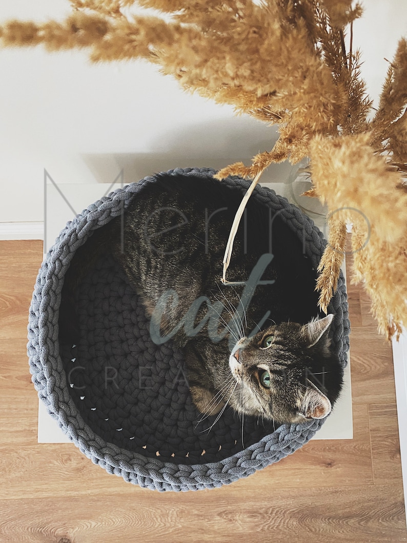 Puede incluir: Una cama para gatos de crochet gris con un gato atigrado descansando dentro. La cama para gatos es redonda y tiene un borde grueso de cuerda.