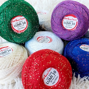 Puede incluir: Una colección de ovillos de lana en varios colores, incluyendo verde, morado, blanco, rojo y azul. Cada ovillo tiene una etiqueta con el nombre de la marca "Ariadna" y el texto "LUMI 10" con el número de color y el peso.