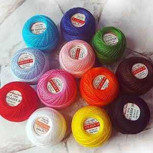 May include: Assortment of 10 balls of embroidery floss in various colors, including red, orange, yellow, green, blue, purple, pink, brown, white, and black. Each ball has a label with the brand name "Atlas" and the text "ATŁASEK 30" in Polish. The balls are arranged in a circle on a white tiled surface.