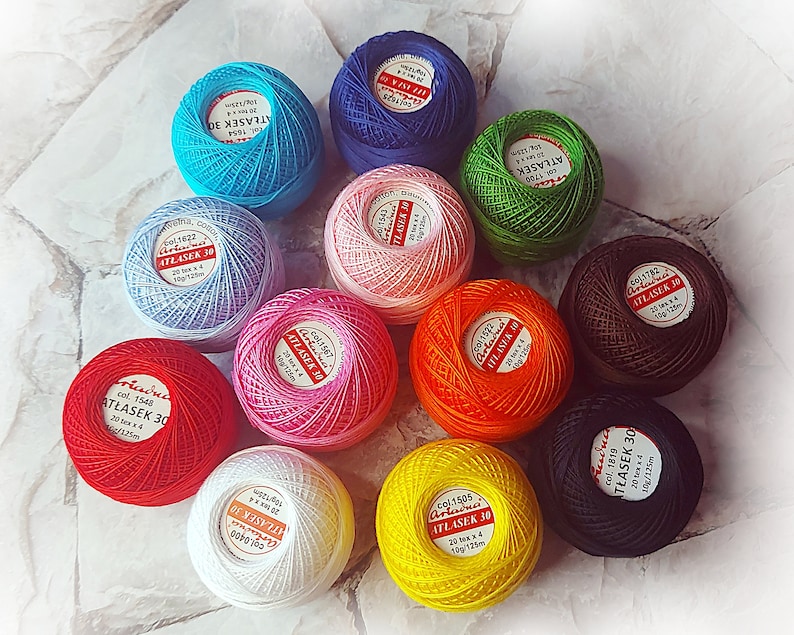May include: Assortment of 10 balls of embroidery floss in various colors, including red, orange, yellow, green, blue, purple, pink, brown, white, and black. Each ball has a label with the brand name "Atlas" and the text "ATŁASEK 30" in Polish. The balls are arranged in a circle on a white tiled surface.