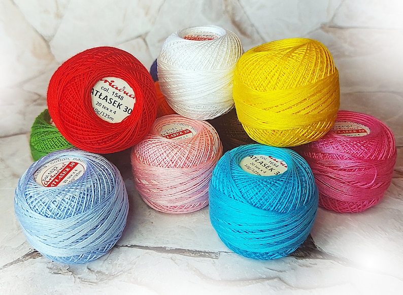 May include: A collection of colorful balls of yarn, each with a label that reads "Criadua Atłasek 30" and "Criadua Atłasek 3". The yarn is available in red, white, yellow, green, light blue, pink, turquoise, and hot pink.
