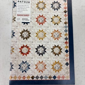 May include: A quilt pattern booklet with a blue cover and a white background featuring a quilt design with a starburst pattern in shades of red, orange, yellow, brown, and blue. The title of the pattern is "Apple Sauce" and the pattern number is "PATO28".