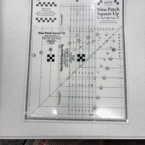 May include: A clear acrylic Nine Patch Square Up quilting ruler tool in its original plastic packaging. The tool features printed black measurement markings, grid lines, and text including Nine Patch Square Up, Part A, and Part B, designed for trimming quilt blocks from 2 to 9 inches.
