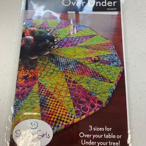 May include: A sewing pattern for a table runner or tree skirt called "Over Under" by Story Girls. The pattern includes three sizes and features a colorful patchwork design.