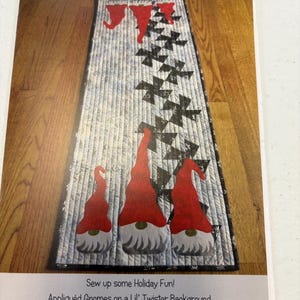 May include: A printed pattern for a quilted table runner with gnome designs. The runner has a light grey background with black and red accents. The text reads "Twister Gnomes" and "Approximately 38 cm W x 99 cm H".