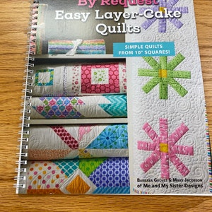 May include: A book titled "By Request Easy Layer-Cake Quilts" with a colorful quilt design on the cover. The book features simple quilts made from 10-inch squares.