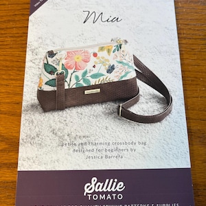 May include: A sewing pattern for a small crossbody bag called "Mia" designed by Jessica Barrera. The pattern features a floral print and a brown leather strap. The pattern is from Sallie Tomato, a company that sells sewing patterns and supplies.