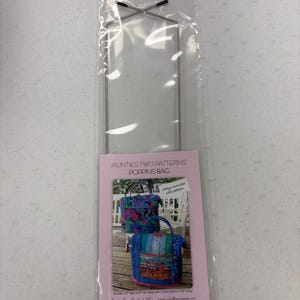 May include: A clear plastic bag containing a metal frame and a pink card. The card features two images of Poppins Bags with floral and striped patterns. Text on the card reads "Aunties Two Patterns Poppins Bag."