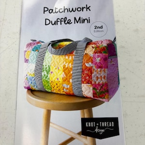 May include: A sewing pattern booklet titled Patchwork Duffle Mini 2nd Edition by Knot + Thread Design. The cover features a colorful quilted duffle bag with black and white striped handles, resting on a wooden stool against a white background.