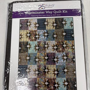 May include: A Westminster Way Quilt Kit featuring One Layer Away by Seth Apter in a 25th Anniversary Free Spirit package. The kit displays a multi-colored quilt design with a repeating star pattern in shades of blue, yellow, brown, and black.