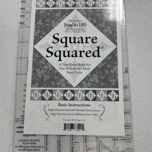 May include: Clear plastic quilting ruler with the text "Square Squared" and "Studio 180 Design". The ruler has measurements from 1" to 6" and includes instructions for right and left-handed use. It is designed for trimming down quilt blocks.