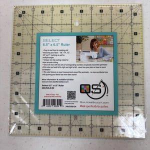 May include: A clear plastic 6.5 inch by 6.5 inch quilting ruler with markings for 1/8, 1/4, 1/2, 3/4 and 1 inch measurements. The ruler has a non-slip coating and features numbers around the perimeter for easy reading. The ruler is made by Quilters Select and is specifically for quilters.