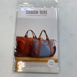 May include: A sewing pattern for a tote bag called "Oxbow Tote" by Noodlehead. The pattern includes two sizes, regular and overnight. The image shows two tote bags, one brown and one blue, with a brown leather handle and strap.